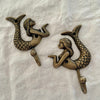 Set of two antique brass mermaid wall hooks shown at angle on linen background revealing hook depth and scaled tail detail
