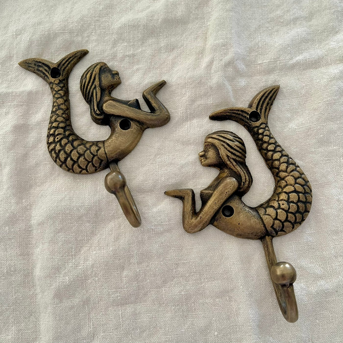 Set of two antique brass mermaid wall hooks shown at angle on linen background revealing hook depth and scaled tail detail
