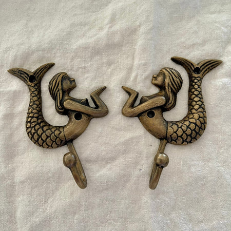 Set of two antique brass mermaid wall hooks facing each other in mirrored display on linen background
