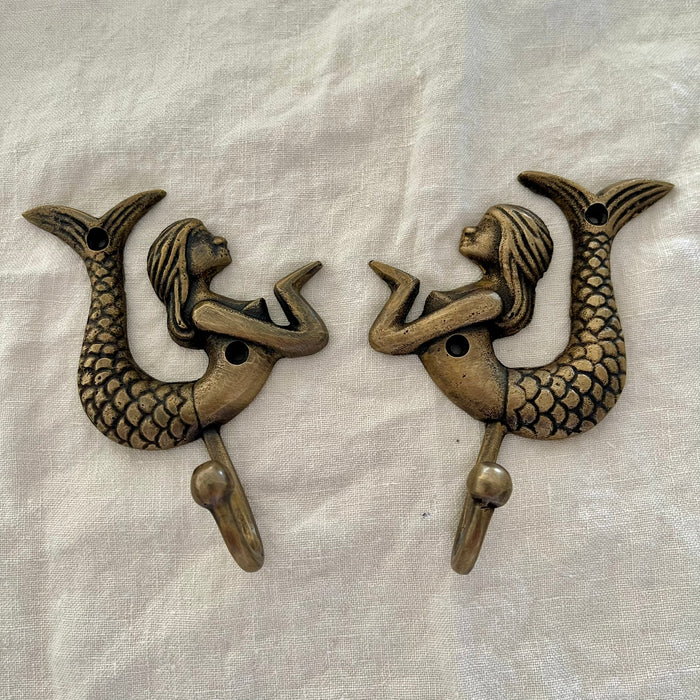 Set of two antique brass mermaid wall hooks facing each other in mirrored display on linen background
