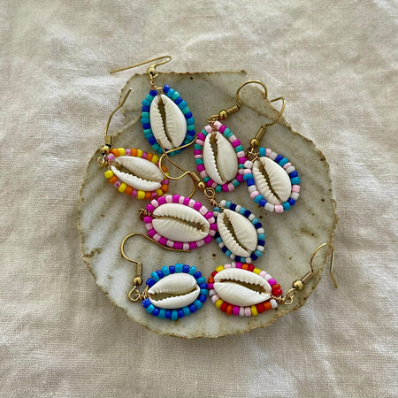 Collection of beaded cowrie shell earrings in multiple colours displayed on a natural shell dish
