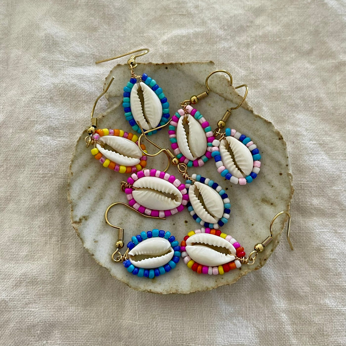 Collection of beaded cowrie shell earrings in multiple colours displayed on a natural shell dish
