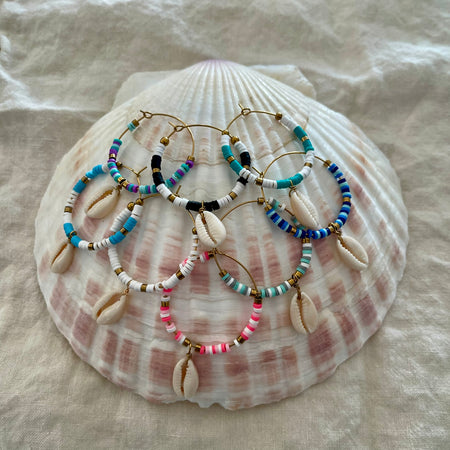 Collection of beaded hoop earrings with cowrie shell drops in multiple colours displayed on a large natural shell
