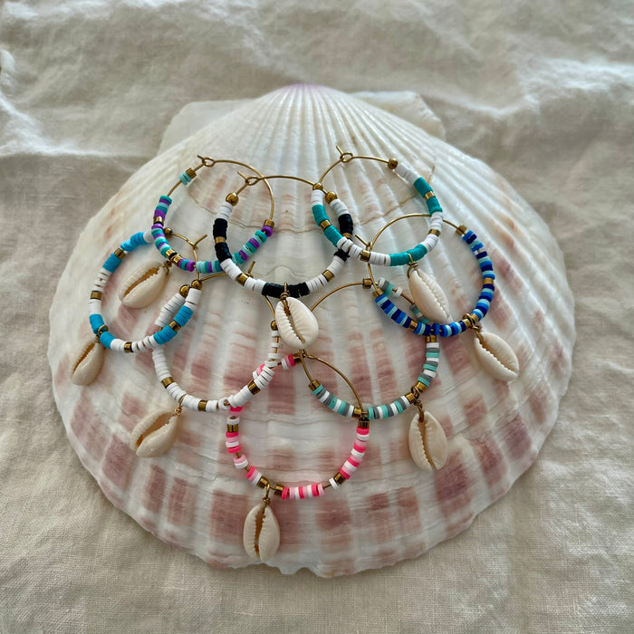Collection of beaded hoop earrings with cowrie shell drops in multiple colours displayed on a large natural shell
