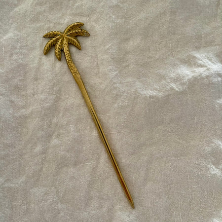 Polished brass palm tree cocktail pick with detailed textured fronds on linen background
