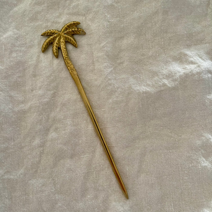 Polished brass palm tree cocktail pick with detailed textured fronds on linen background
