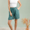 100% linen Bermuda shorts in duck green front view with drawstring waist and pom-pom trim hem worn by model with dried floral arrangement
