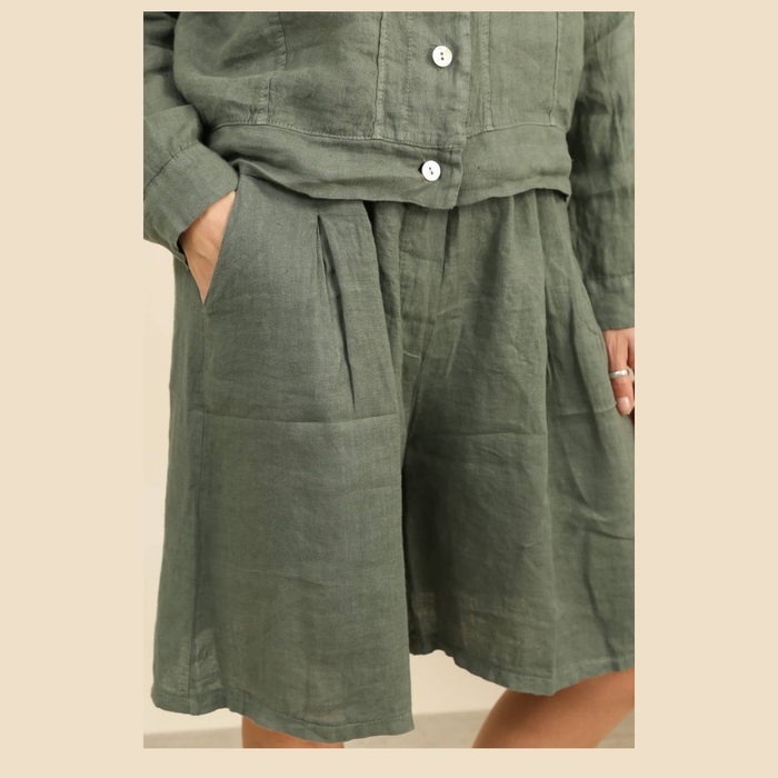 100% linen wide-leg Bermuda shorts in duck green side view worn by model with matching linen jacket and tropical plant
