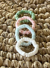 Four pastel shell disc stretch rings in pink, green, blue and white, overlapping on a natural seagrass surface

