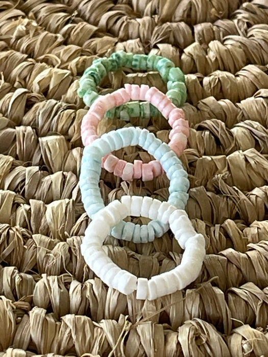 Four pastel shell disc stretch rings in pink, green, blue and white, overlapping on a natural seagrass surface
