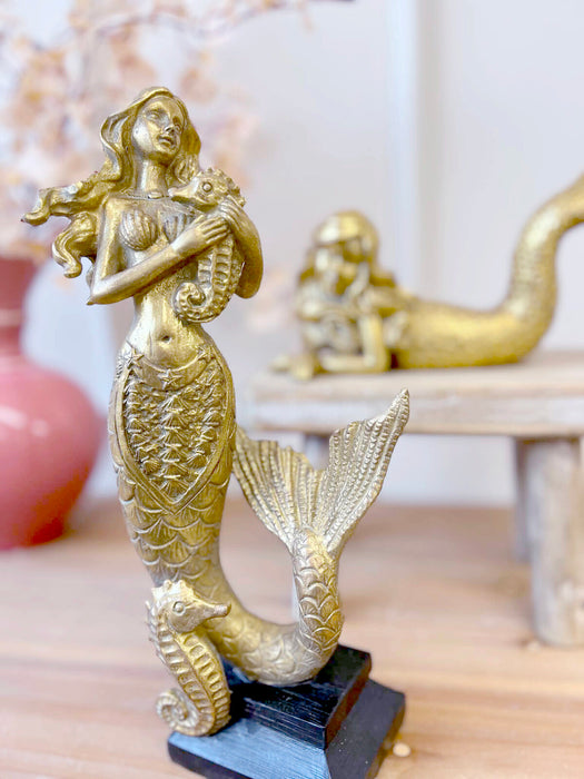 Gold resin standing mermaid sculpture styled on timber shelf with laying mermaid and pink vase
