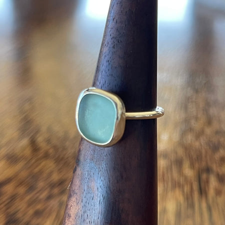 Sterling silver blue seaglass ring on wooden ring cone, side view showing freeform aqua seaglass in simple bezel setting with slim hammered band
