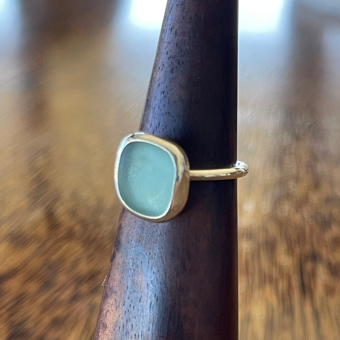 Sterling silver blue seaglass ring on wooden ring cone, side view showing freeform aqua seaglass in simple bezel setting with slim hammered band
