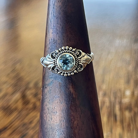 Sterling silver blue topaz detailed ring on wooden ring cone, front view showing round-cut blue topaz in rope-twist bezel with scrollwork and bead surround
