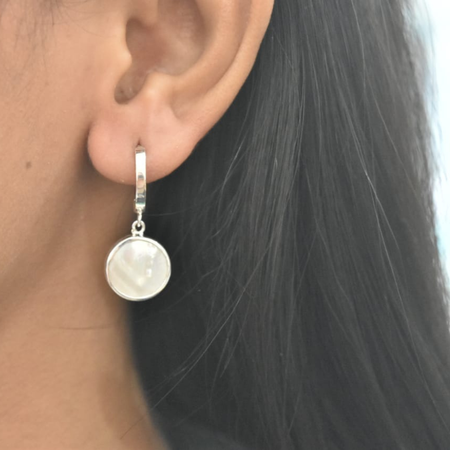 Sterling silver mother of pearl disc drop earrings worn on ear, showing round iridescent mother of pearl pendant suspended from a sleek silver huggie hoop
